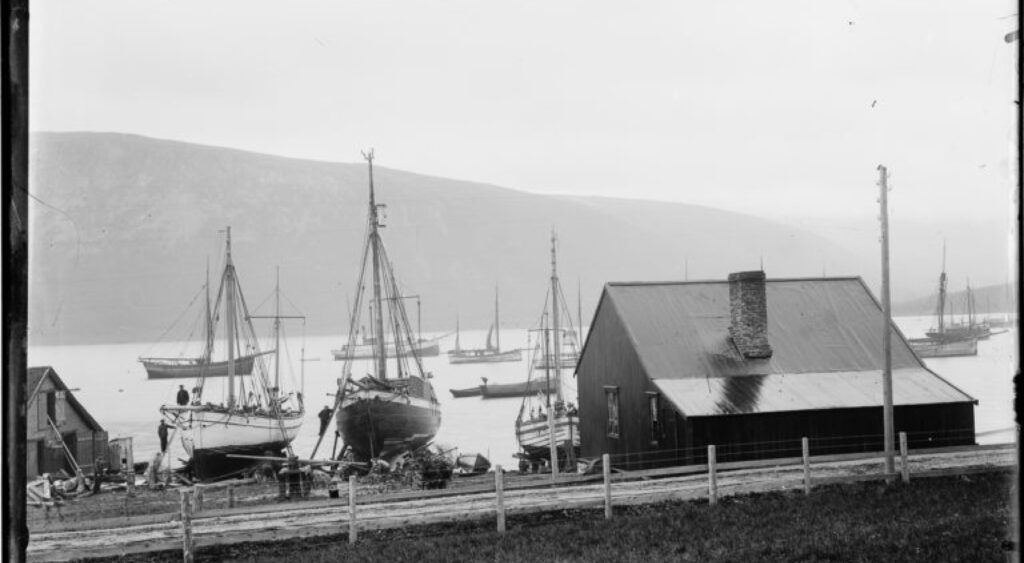 Et gammelt sort hvitt bilde av en bygning og båter ved sjøkanten